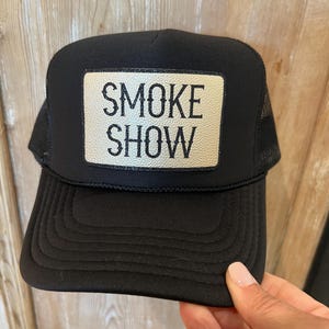 Puede incluir: Gorra de camionero negra con parte trasera de malla y visera curva. La gorra presenta un parche rectangular con las palabras "SMOKE SHOW" en letras blancas y en negrita. Un accesorio cl&aacute;sico.