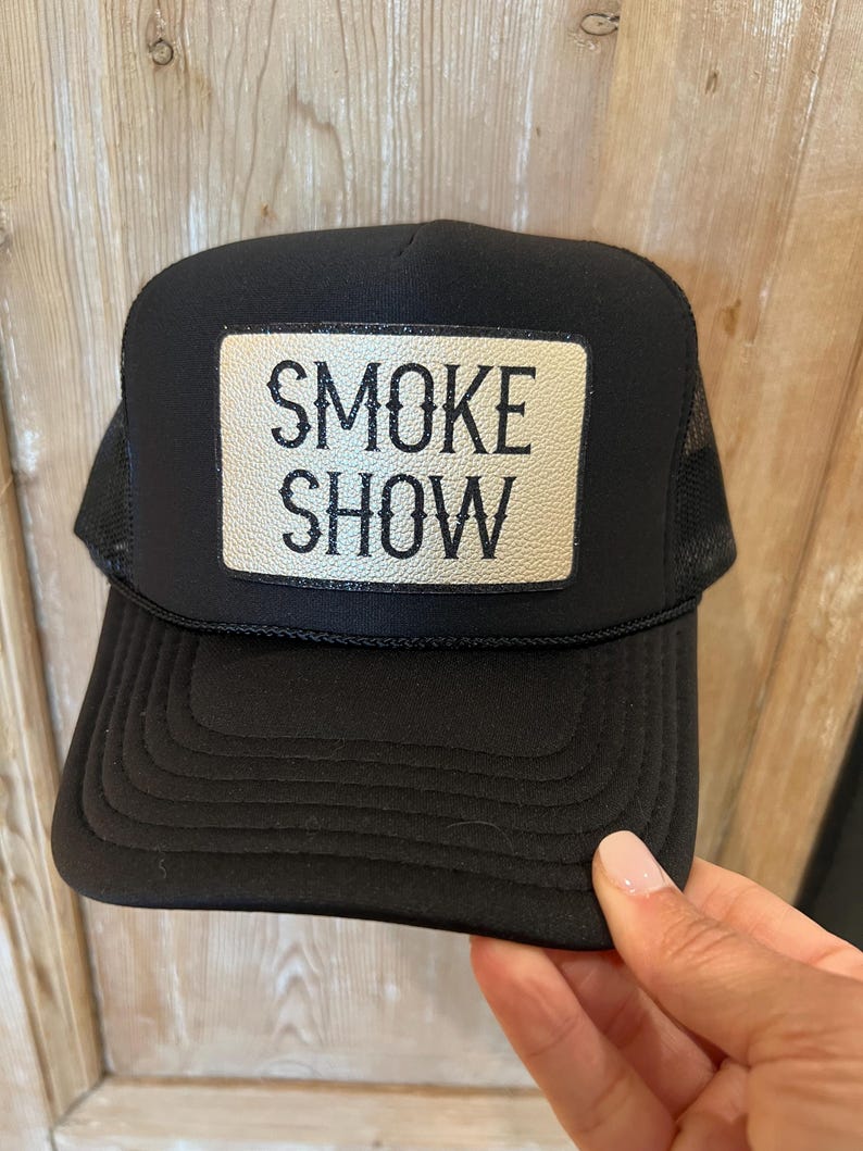 Puede incluir: Gorra de camionero negra con parte trasera de malla y visera curva. La gorra presenta un parche rectangular con las palabras "SMOKE SHOW" en letras blancas y en negrita. Un accesorio cl&aacute;sico.