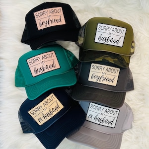 May include: Six trucker hats with different colored mesh backs and embroidered patches. The patches have the text "Sorry About My" followed by "boyfriend", "husband", "ex-husband".