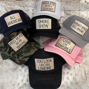 Puede incluir: Una colecci&oacute;n de gorras de camionero en varios colores, incluyendo azul marino, negro, gris, blanco, rosa y camuflaje. Cada gorra presenta un parche rectangular con textos como "All American" y "Long Live Cowgirls."