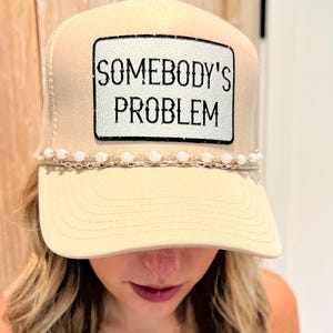 Puede incluir: Gorra de camionero beige con un parche blanco que dice "SOMEBODY'S PROBLEM" en texto negro. El sombrero tiene un detalle de perlas y cadena a lo largo del borde. La gorra tiene una parte trasera de malla y una visera curva.