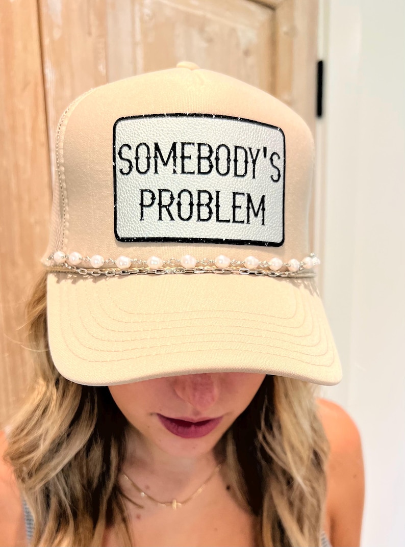 Puede incluir: Gorra de camionero beige con un parche blanco que dice "SOMEBODY'S PROBLEM" en texto negro. El sombrero tiene un detalle de perlas y cadena a lo largo del borde. La gorra tiene una parte trasera de malla y una visera curva.
