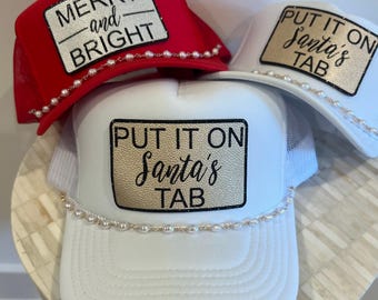 May include: Three trucker hats with holiday-themed text. One red hat says "MERRY and BRIGHT" and is trimmed with pearls. Two white hats say "PUT IT ON Santa's TAB" and are also trimmed with pearls. The hats have a mesh back.