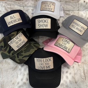 Puede incluir: Una colecci&oacute;n de gorras de camionero en varios colores, incluyendo azul marino, negro, gris, blanco, rosa y camuflaje. Cada gorra presenta un parche rectangular con texto como "All American", "Smoke Show" y "Long Live Cowgirls."