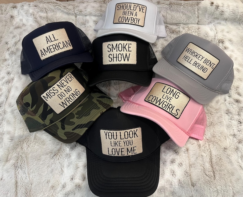 Puede incluir: Una colecci&oacute;n de gorras de camionero en varios colores, incluyendo azul marino, negro, gris, blanco, rosa y camuflaje. Cada gorra presenta un parche rectangular con texto como "All American", "Smoke Show" y "Long Live Cowgirls."