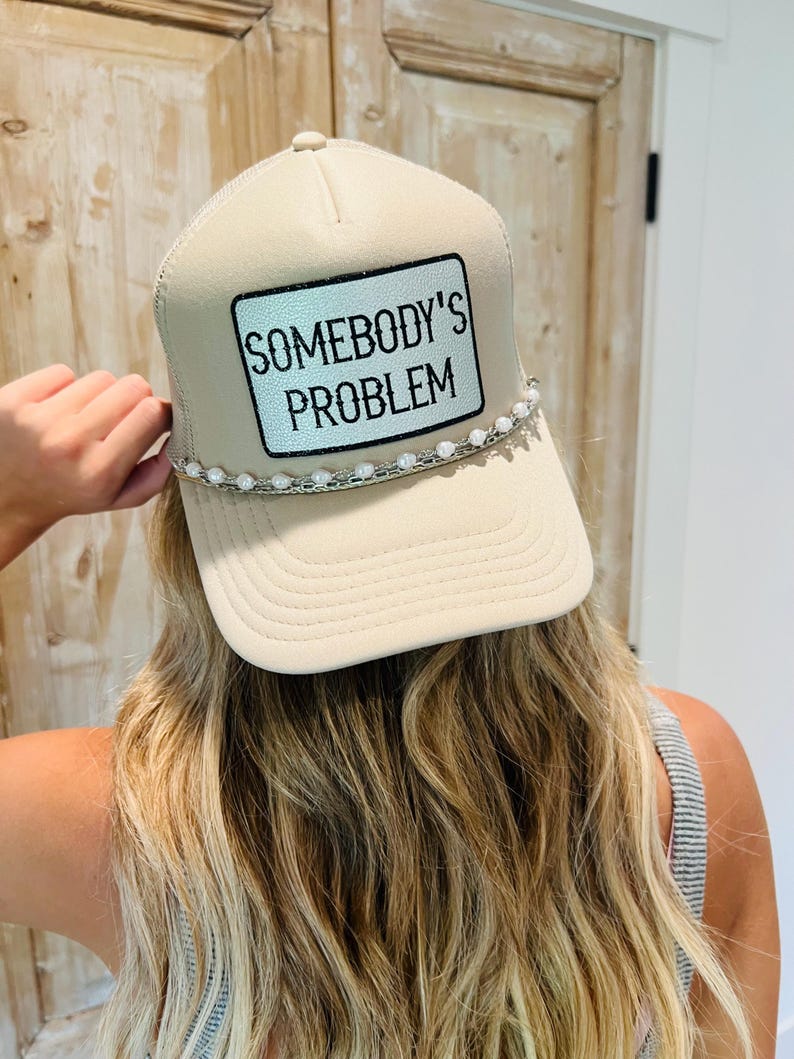 Puede incluir: Gorra de camionero beige con parte trasera de malla y visera curva. La parte delantera presenta un parche rectangular con el texto "SOMEBODY'S PROBLEM" en negro. Una hilera de cuentas blancas decora la visera.