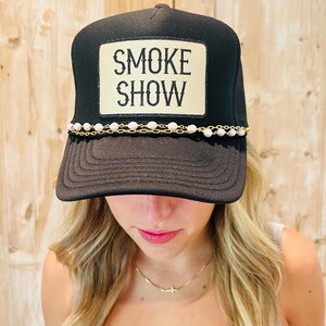 Puede incluir: Gorra de camionero negra con parte trasera de malla y visera curva. La parte delantera presenta un parche rectangular con las palabras "SMOKE SHOW" en negro. Una cadena dorada con cuentas blancas adorna la visera.