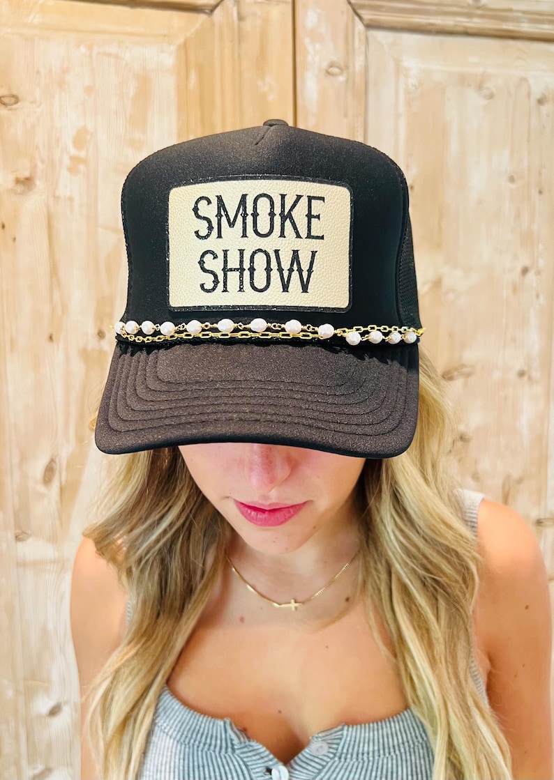 Puede incluir: Gorra de camionero negra con parte trasera de malla y visera curva. La parte delantera presenta un parche rectangular con las palabras "SMOKE SHOW" en negro. Una cadena dorada con cuentas blancas adorna la visera.