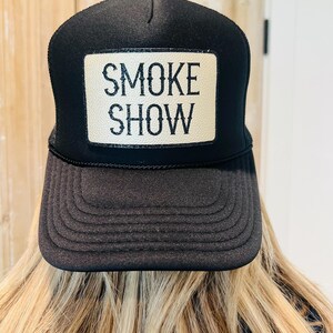 Puede incluir: Gorra de camionero negra con un parche rectangular que dice "SMOKE SHOW" en letras blancas. La gorra tiene una visera curva y una parte trasera de malla. La gorra la lleva una persona con pelo rubio.