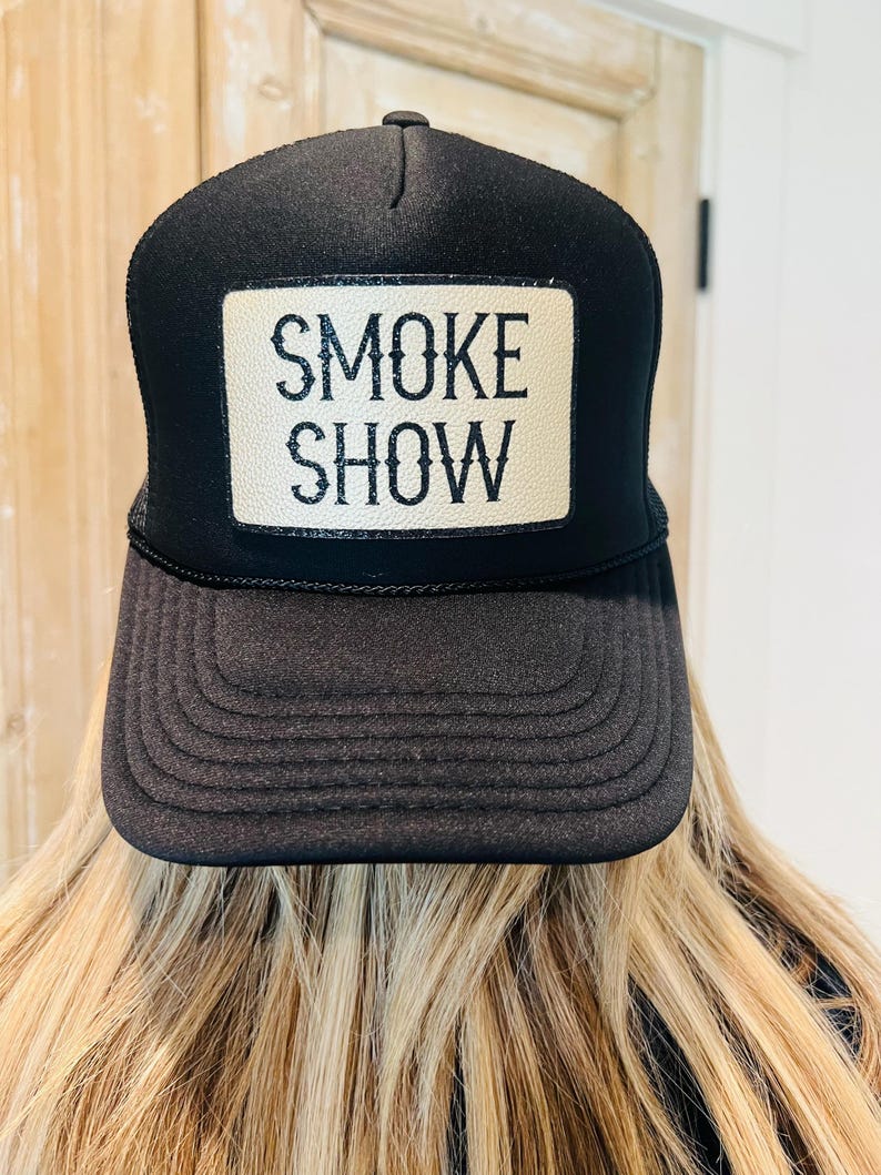 Puede incluir: Gorra de camionero negra con un parche rectangular que dice "SMOKE SHOW" en letras blancas. La gorra tiene una visera curva y una parte trasera de malla. La gorra la lleva una persona con pelo rubio.