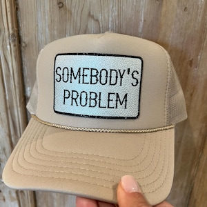 Puede incluir: Gorra trucker beige con parte trasera de malla y visera curva. El panel frontal presenta un parche rectangular con el texto "SOMEBODY'S PROBLEM" en letras negras. Un cord&oacute;n dorado adorna la visera.