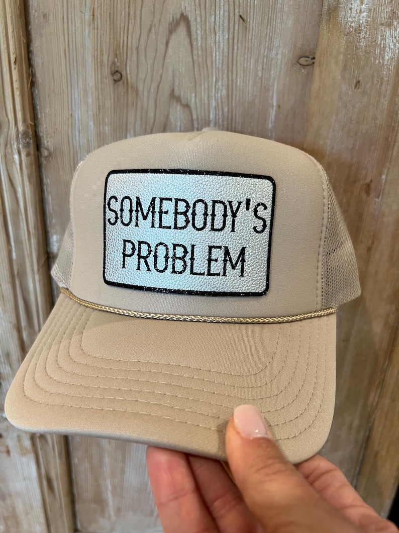 Puede incluir: Gorra trucker beige con parte trasera de malla y visera curva. El panel frontal presenta un parche rectangular con el texto "SOMEBODY'S PROBLEM" en letras negras. Un cord&oacute;n dorado adorna la visera.