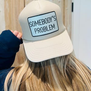 Puede incluir: Gorra de camionero beige con un parche blanco que dice "SOMEBODY'S PROBLEM" en texto negro. La gorra tiene una visera curva y una parte trasera de malla. La persona que lleva la gorra tiene el pelo largo y rubio.