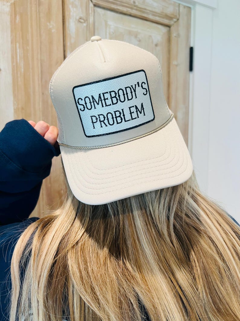 Puede incluir: Gorra de camionero beige con un parche blanco que dice "SOMEBODY'S PROBLEM" en texto negro. La gorra tiene una visera curva y una parte trasera de malla. La persona que lleva la gorra tiene el pelo largo y rubio.