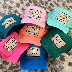 May include: A collection of seven colorful trucker hats featuring mesh backs and rectangular patches with black text. The hats are pink, teal, light blue, orange, hot pink, dark green, and blue, with phrases like hello sunshine, sunkissed, tropic like it's hot, boujee on the beach, chasing sunsets, vacay mode, and beach girl.