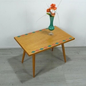 Pode incluir: Uma mesa de centro de estilo moderno de meados do século com acabamento em madeira clara e pernas anguladas. A mesa apresenta uma borda decorativa de formas de diamante turquesa e coral alternadas. Um vaso turquesa com flores laranja está sobre a mesa.