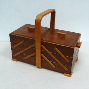 Può includere: Un portagioie vintage in legno con manico marrone chiaro e corpo marrone scuro. La scatola ha due scomparti con coperchi e sezioni angolate a forma di cassetto. La scatola è in legno e ha un design classico.