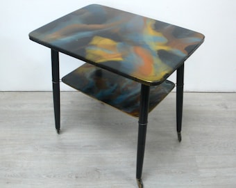 May include: A two-tier side table featuring a rectangular top and a smaller rectangular lower shelf, both decorated with a swirling abstract pattern of blue, orange, and yellow colors. The table has four black tapered legs with small casters.