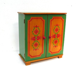 May include: A small wooden cabinet with two doors, featuring a green frame and light wood panels. Each door is decorated with a red and green floral pattern and a small red knob. The cabinet stands on four short, rectangular wooden feet.