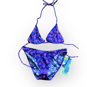 Puede incluir: Un bikini de estampado floral morado y rosa con una parte superior triangular y una parte inferior de corte alto. El bikini es de la marca "Malibu" y tiene una etiqueta azul.