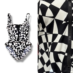 May include: A black and white geometric patterned swimsuit with a front zipper. The one-piece swimsuit features a bold, abstract design. The straps are thin and adjustable. The fabric has a repeating pattern of black and white shapes.