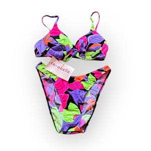 May include: A two-piece swimsuit with a bright floral print in shades of pink, green, purple, and black. The top is a halter style with a twist detail in the front. The bottom is a high-cut style with a cheeky fit. The swimsuit is labelled "carabella collections".