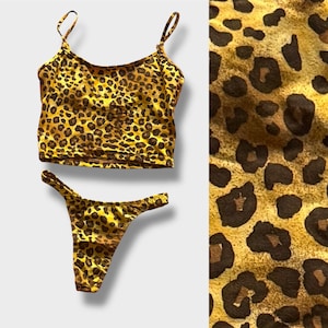 Könnte beinhalten: Ein zweiteiliges Set mit Leopardenmuster. Das Set besteht aus einem Camisole-Top mit dünnen Trägern und einem Höschen im String-Stil. Der Stoff hat ein gelb-braunes Farbschema mit dunkelbraunen Flecken.
