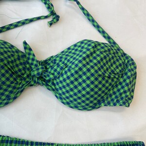 May include: A two-piece swimsuit set in a green and navy blue gingham pattern. The top is a halter-style bikini with a bow detail, and the bottoms are pleated. The fabric has a soft, textured appearance.