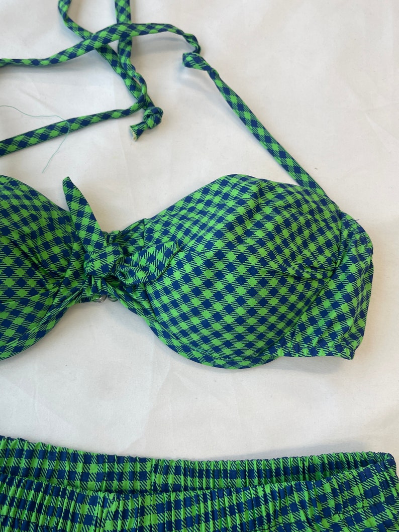 May include: A two-piece swimsuit set in a green and navy blue gingham pattern. The top is a halter-style bikini with a bow detail, and the bottoms are pleated. The fabric has a soft, textured appearance.