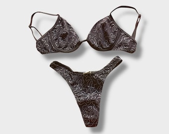 May include: A black lingerie set featuring a bra and a thong. The set is adorned with a silver, textured pattern. The bra has underwire support and adjustable straps. The thong has a high-cut design. The set is displayed against a white background.