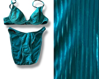 Puede incluir: Conjunto de bikini de color turquesa, confeccionado en tejido acanalado. La parte superior tiene tirantes ajustables y forma triangular. La parte inferior a juego tiene un corte de pierna alto. El conjunto se muestra sobre un fondo blanco.