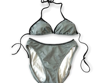 Può includere: Un bikini grigio con bordi neri. La parte superiore è di stile triangolare con spalline regolabili. La parte inferiore è di stile a taglio alto con un bordo nero intorno alla vita.