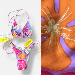 May include: A two-piece bikini set with a floral pattern in shades of purple, pink, and orange. The bikini top has a halter neck design, and the bottom is a thong style. The fabric features a close-up of an orange flower with gold accents.