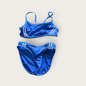 May include: A two-piece swimsuit in royal blue featuring thin shoulder straps and light blue piping trim. The top and bottom pieces are accented with two vertical white stripes on each side, creating a coordinated athletic look for swimming.