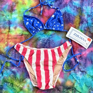 May include: A two-piece bikini set featuring a blue triangle top with white stars and matching bottoms with red and white vertical stripes. The set includes thin blue tie strings and a white tag labeled ZULIANA with size M printed on it.