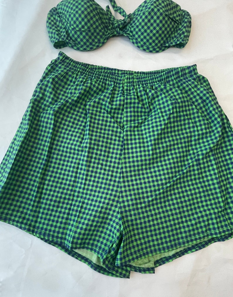 May include: A two-piece swimsuit set in a green and navy blue gingham pattern. The top is a halter-style bikini, and the bottoms are high-waisted shorts. The fabric has a soft, textured appearance. The set is designed for swimwear.