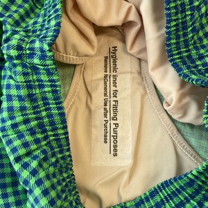 May include: Close-up of a pair of green and blue checkered swim trunks. The interior lining is a light beige color. A white label reads "Hygienic liner for Fitting Purposes. Remove after Purchase."