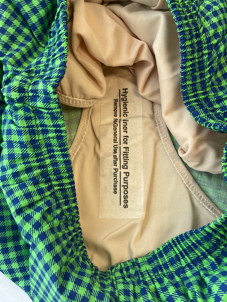 May include: Close-up of a pair of green and blue checkered swim trunks. The interior lining is a light beige color. A white label reads "Hygienic liner for Fitting Purposes. Remove after Purchase."
