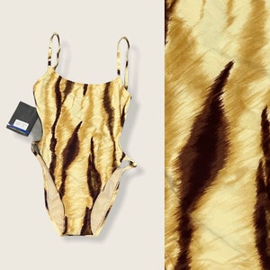 Puede incluir: Un traje de baño marrón y beige con estampado de rayas de tigre. El traje de baño tiene un escote redondo y tirantes finos. El traje de baño tiene recortes en los lados.