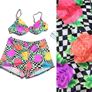 May include: A two-piece swimsuit set featuring a bikini top with thin straps and high-waisted shorts. The fabric displays a black and white checkered pattern overlaid with vibrant, large floral prints in shades of neon pink, orange, yellow, and blue.