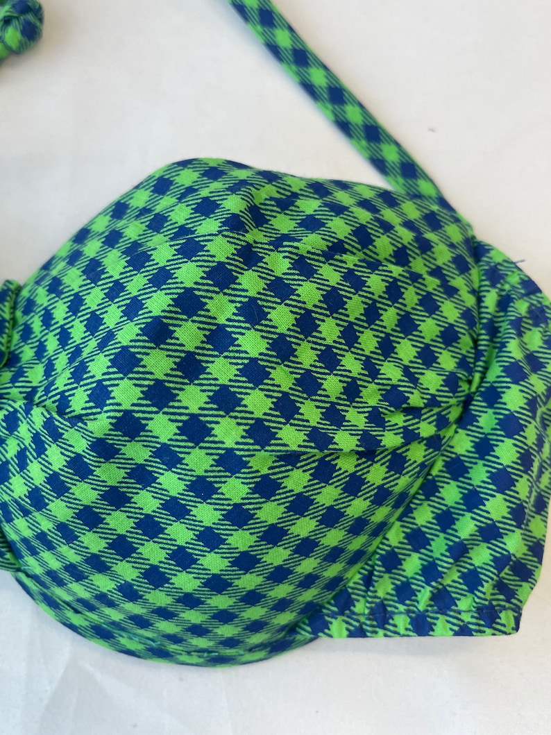 May include: A green and blue checkered bikini top. The top features a classic bra-style design with adjustable straps. The fabric has a repeating diamond pattern in shades of green and blue. The bikini top is made of a soft, lightweight material.