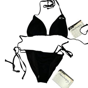 Könnte beinhalten: Schwarzes Bikini-Set mit Triangel-Oberteil und Bindebändern. Das Oberteil hat ein kleines weißes Logo auf einer der Cups. Die passenden Unterteile haben Bindebänder. Enthält Etiketten mit dem Text "Perform Beautifully".