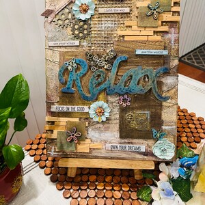 Op de afbeelding: Een mixed media canvas met een bruine achtergrond en het woord "Relax" in blauw en groen in het midden. Het canvas is versierd met bloemen, tandwielen, vlinders en verschillende texturen. Het canvas staat op een houten ezel en heeft de volgende tekst: "light the way", "use your wings", "see the world", "FOCUS ON THE GOOD" en "OWN YOUR DREAMS".