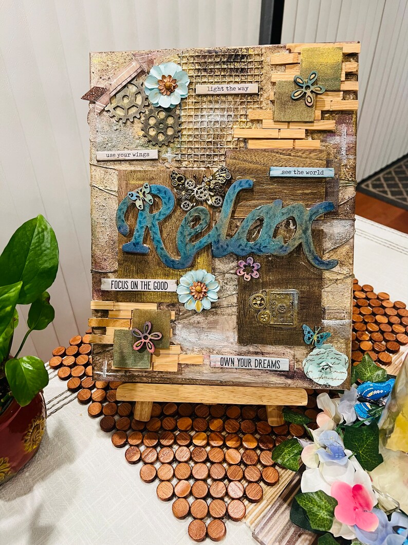 Op de afbeelding: Een mixed media canvas met een bruine achtergrond en het woord "Relax" in blauw en groen in het midden. Het canvas is versierd met bloemen, tandwielen, vlinders en verschillende texturen. Het canvas staat op een houten ezel en heeft de volgende tekst: "light the way", "use your wings", "see the world", "FOCUS ON THE GOOD" en "OWN YOUR DREAMS".