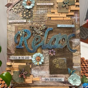 Op de afbeelding: Een houten canvas met een mixed media collage met het woord "Relax" in blauwe en bruine letters. De collage bevat verschillende texturen, kleuren en versieringen, waaronder bloemen, tandwielen, vlinders en inspirerende citaten zoals "use your wings", "light the way", "see the world", "focus on the good" en "own your dreams".