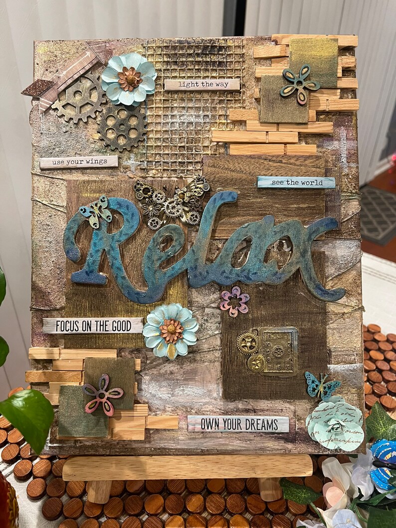 Op de afbeelding: Een houten canvas met een mixed media collage met het woord "Relax" in blauwe en bruine letters. De collage bevat verschillende texturen, kleuren en versieringen, waaronder bloemen, tandwielen, vlinders en inspirerende citaten zoals "use your wings", "light the way", "see the world", "focus on the good" en "own your dreams".