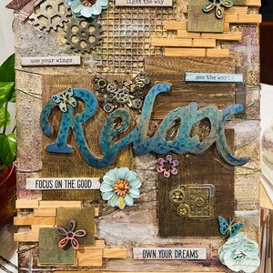 Op de afbeelding: Een houten canvas met een mixed media collage met het woord "Relax" in blauwe en groene letters. De collage bevat verschillende versieringen zoals tandwielen, vlinders, bloemen en houten blokken met inspirerende citaten zoals "use your wings", "light the way", "see the world", "focus on the good" en "own your dreams".