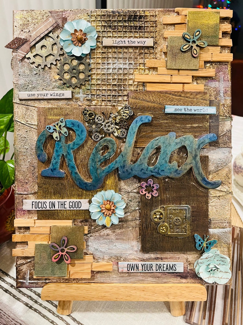 Op de afbeelding: Een houten canvas met een mixed media collage met het woord "Relax" in blauwe en groene letters. De collage bevat verschillende versieringen zoals tandwielen, vlinders, bloemen en houten blokken met inspirerende citaten zoals "use your wings", "light the way", "see the world", "focus on the good" en "own your dreams".