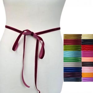 Op de afbeelding: Een diepbordeauxrood fluwelen lint gestrikt rond een witte etalagepop-torso. Rechts is een collectie fluwelen lintspoelen in een raster weergegeven, met een verscheidenheid aan kleuren waaronder goud, rood, groen, blauw, zwart en roze.