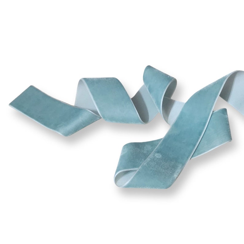 K&ouml;nnte beinhalten: Ein Band aus teal-farbenem Samtband, das sich vor einem wei&szlig;en Hintergrund kr&auml;uselt und wellt. Die weiche Textur und Farbe des Bandes werden hervorgehoben und zeigen sein Potenzial f&uuml;r Bastelarbeiten, Geschenkverpackungen oder dekorative Zwecke.